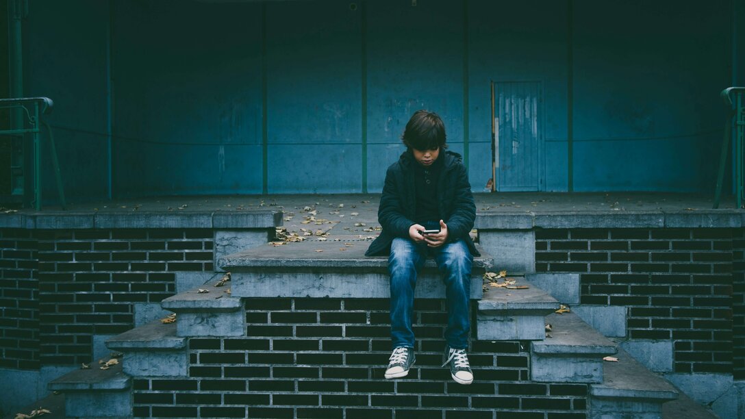 Ein Junge sitzt allein auf einer Treppe vor einer Lagerhalle und schaut auf sein Handy | © Gaelle Marcel, Unsplash